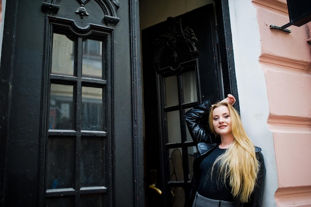 Blonde fashionable girl in long black leather coat posed against wooden door at old house