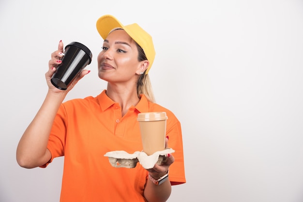 Blonde female courier drinking cup of coffee on white wall. 