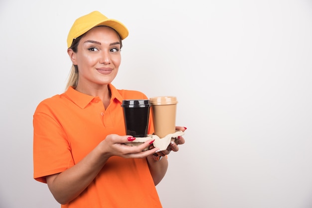 Blonde female courier holding two cups of coffees looking sideways on white wall. 