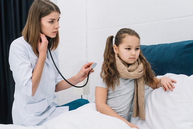 Белокурая женщина-врач осматривает девушку, сидящую на кровати в больнице