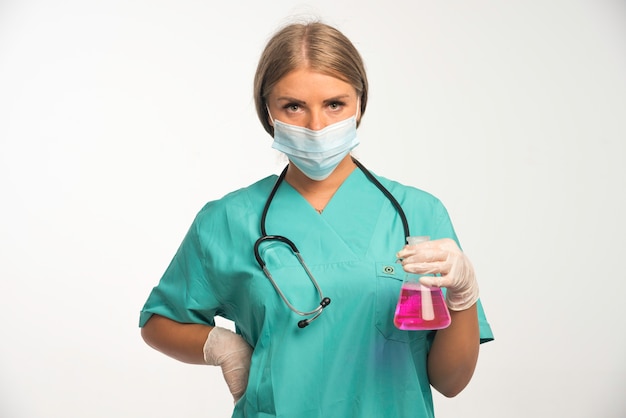 Блондинка женщина-врач в синей форме со стетоскопом на шее в маске для лица и с химической колбой.