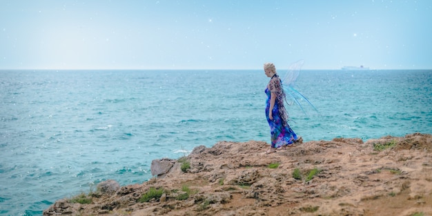 Blonde female dressed as a fairy standing on the shore surrounded by the sea under a blue sky
