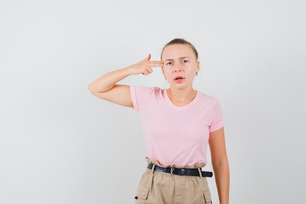 Blonde female making suicide gesture in t-shirt, pants and looking serious , front view.