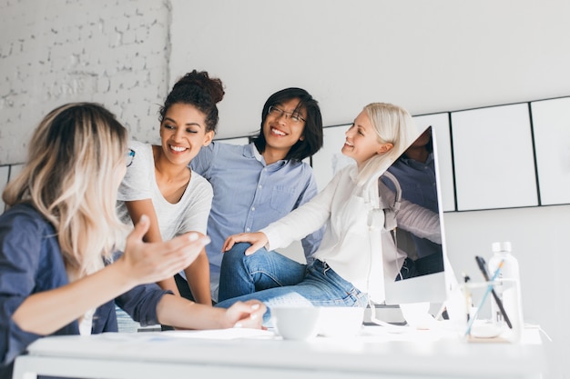 Blonde female secretary telling funny story to laughing colleagues. Indoor portrait of smiling asian office worker listening at fair-haired friend, standing beside computer.