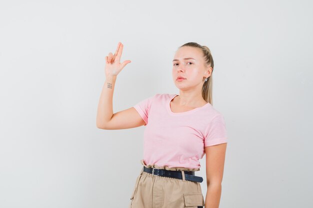 Blonde female showing gun gesture in t-shirt, pants and looking strict. front view.