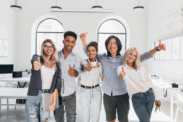 Blonde female student in ripped jeans holding laptop and laughing after hard test. Asian guy in glasses waving hands, celebrating end of exams and beginning of vacation.