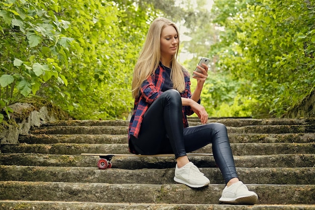Блондинка с помощью смартфона сидит на лонгборде на ступеньках.