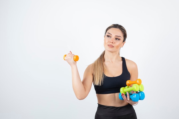 Blonde fit woman in black top standing and holding colorful dumbbells.  