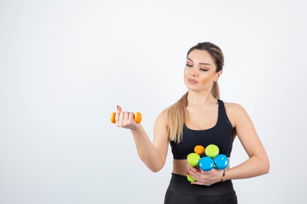 Blonde fit woman in black top standing and holding colorful dumbbells.  