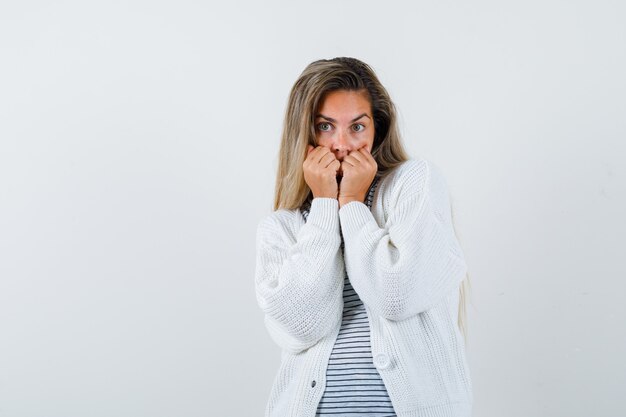 Blonde girl biting fists emotionally in striped t-shirt, white cardigan and jean pants and looking timid. front view.