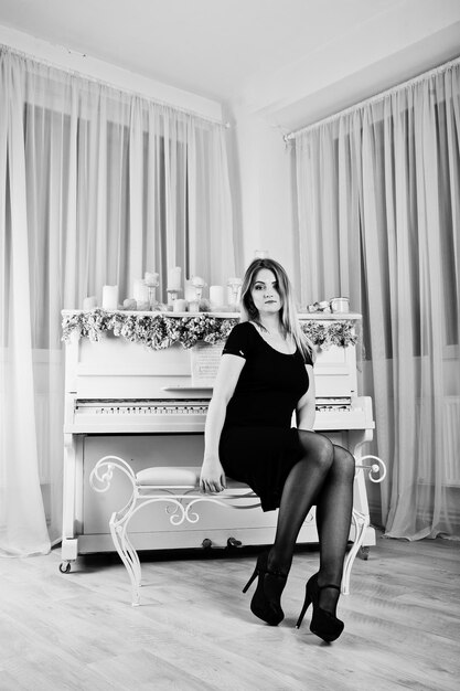 白い部屋でクリスマスキャンドルの装飾とピアノの近くでポーズをとった黒いドレスのブロンドの女の子