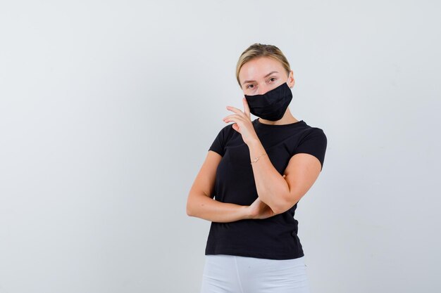 Blonde girl in black t-shirt, white pants, black mask leaning isolated
