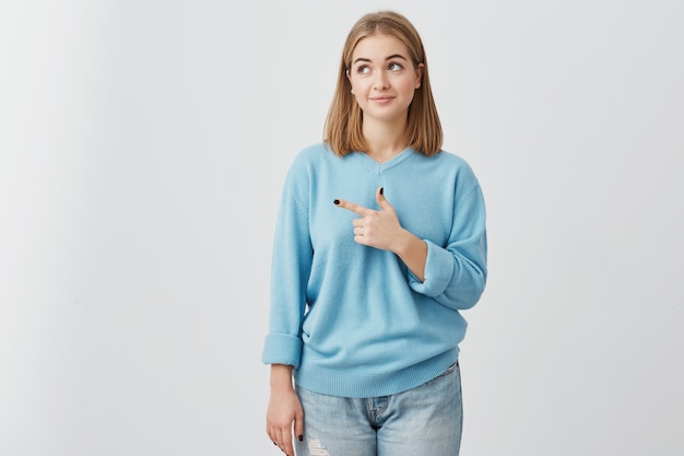 Белокурая девушка в голубом свитере смотря с ее темными глазами в сторону указывать с указательным пальцем на рекламу космоса экземпляра что-то. Женщина представляя против серой стены с космосом экземпляра для текста или продвижения