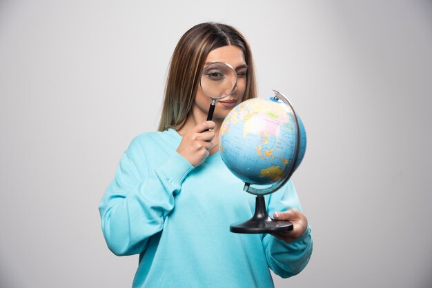 Blonde girl in blue sweatshirt holding a globe and looking for a destination with magnifier.