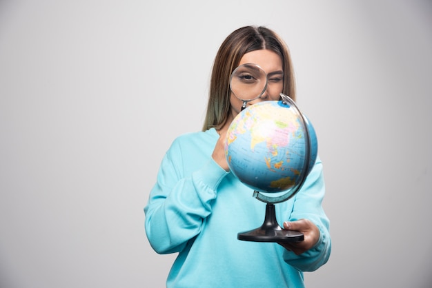 Blonde girl in blue sweatshirt holding a globe and looking for a destination with magnifier.