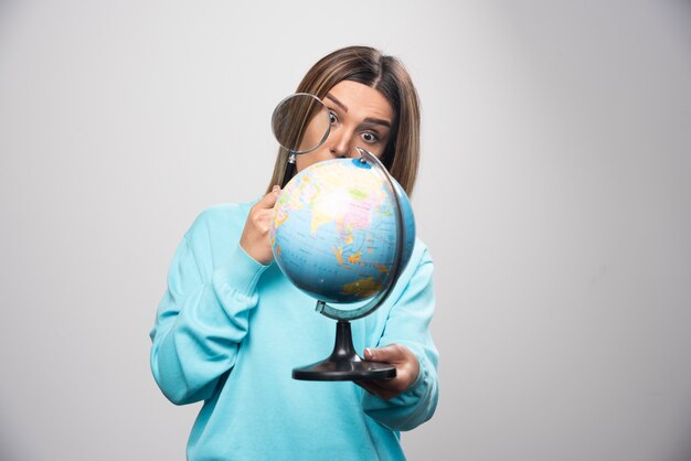 Blonde girl in blue sweatshirt holding a globe and looking for a destination with magnifier.