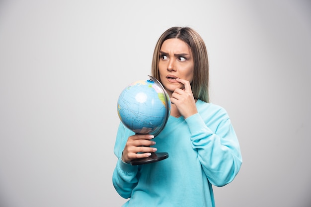 Blonde girl in blue sweatshirt holding a globe, thinking carefully and trying to remember