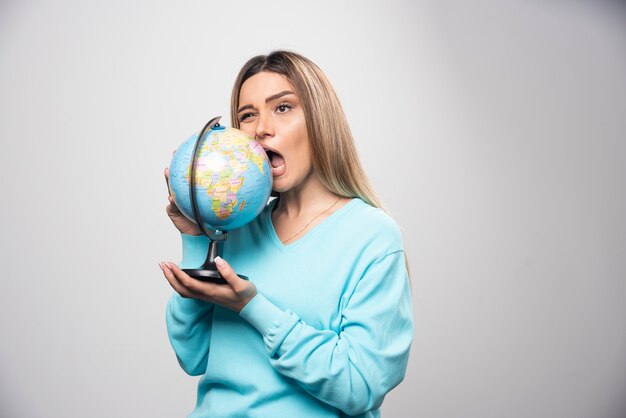 Blonde girl in blue sweatshirt holds a globe and bites it.