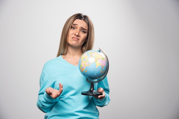 Blonde girl in blue sweatshirt holds a globe and looks uncertain and confused.