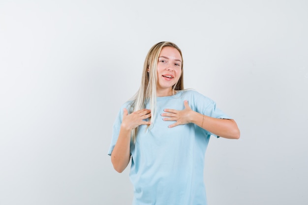 Blonde girl in blue t-shirt holding hands over chest and looking cheery , front view.