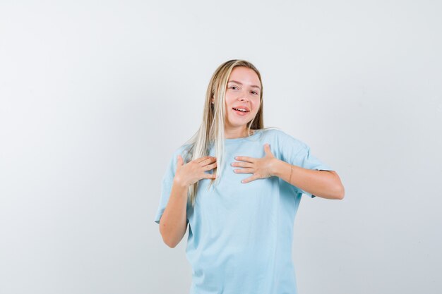 胸に手をかざして陽気に見える青いTシャツのブロンドの女の子、正面図。