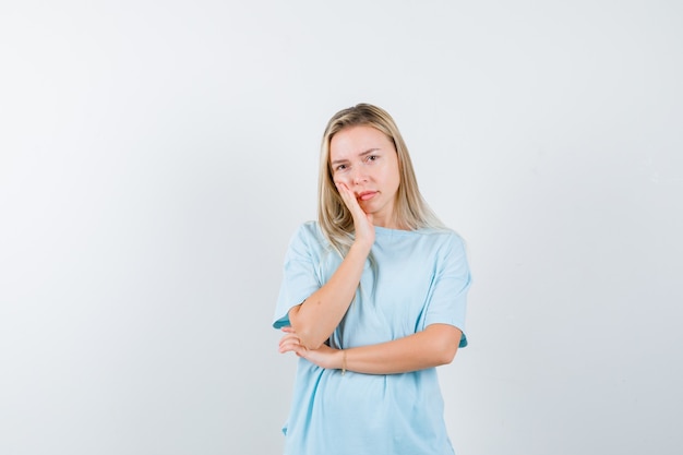 Blonde girl in blue t-shirt leaning cheek on palm, holding hand under elbow and looking pretty , front view.