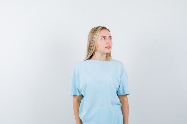 Blonde girl in blue t-shirt looking away while posing at camera and looking pretty , front view.