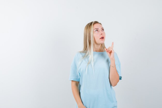 Blonde girl in blue t-shirt pointing up with index finger, looking upward and looking pensive , front view.