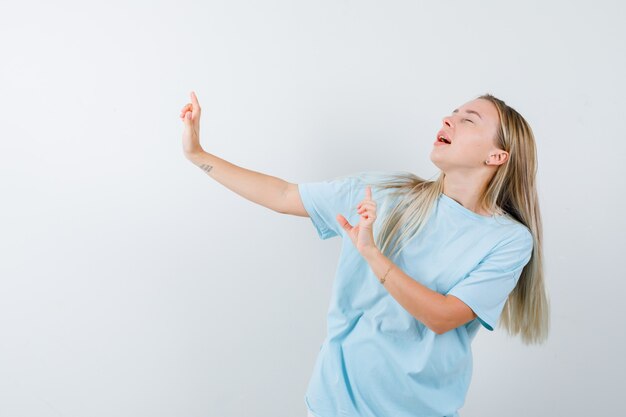 Blonde girl in blue t-shirt showing gun gestures, keeping eyes closed and looking cheery , front view.