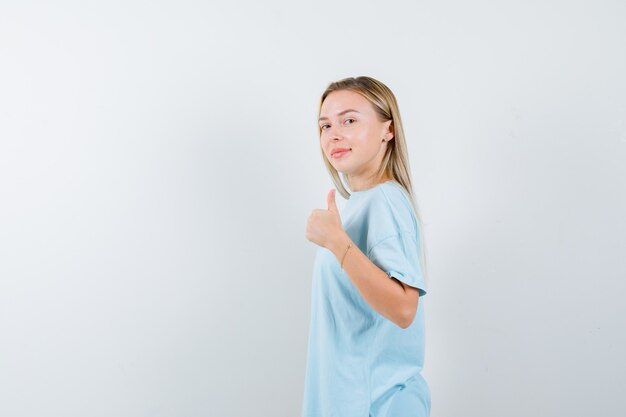 親指を上に向けて、肩越しに見て、自信を持って、正面図を示す青いTシャツのブロンドの女の子。