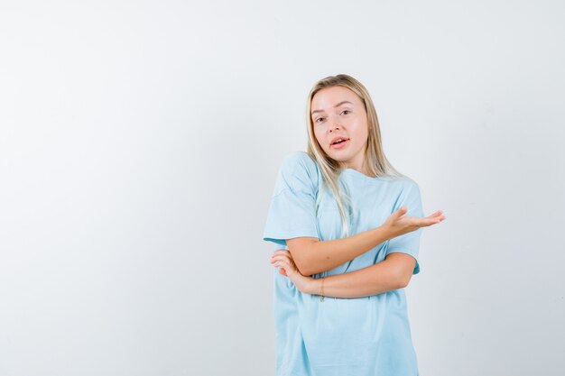 Blonde girl in blue t-shirt shrugging shoulders, showing helpless gesture and looking puzzled , front view.