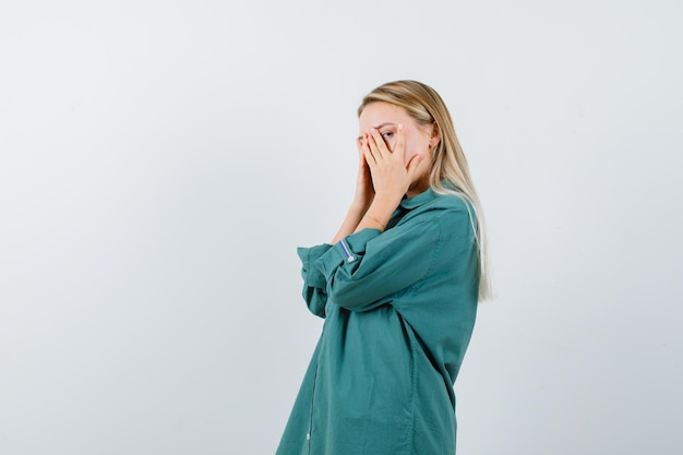 Blonde girl covering face with hands, looking through fingers in green blouse and looking serious.