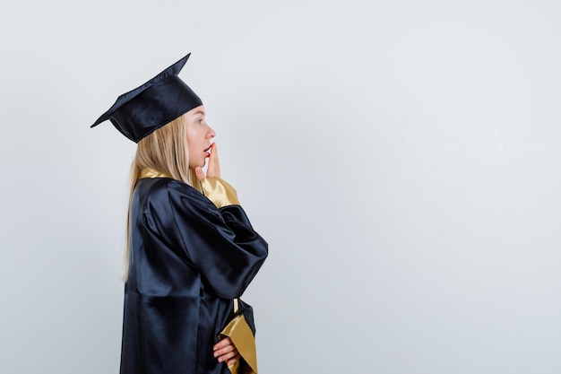 Blonde girl covering mouth with hand in graduation gown and cap and looking surprised.