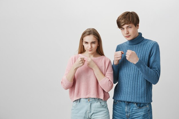 Blonde girl and fair-haired guy standing indoors with fists bunched and determined face expressions.