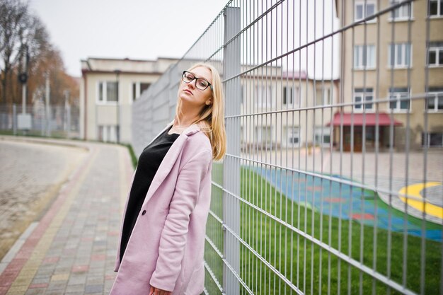 Blonde girl at glasses and pink coat black tunic and handbag posed against fence at street