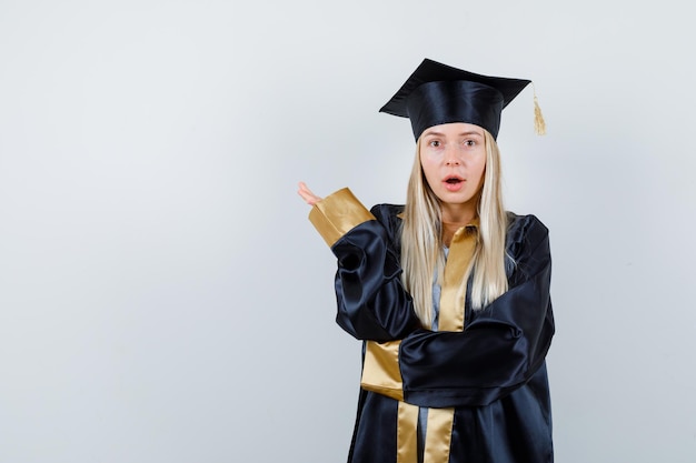 手を上げて、口を開けて、困惑しているように見える大学院の制服を着たブロンドの女の子