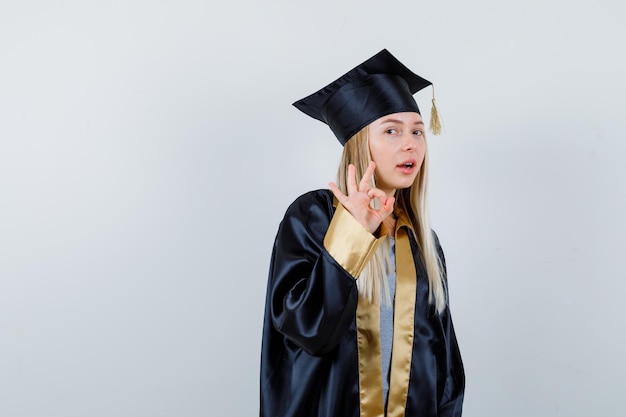 大丈夫なジェスチャーを示し、自信を持って見える大学院の制服を着たブロンドの女の子