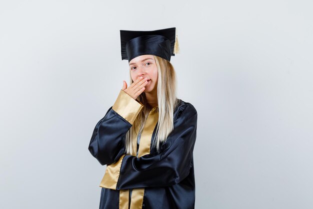 Ragazza bionda in abito da laurea e berretto che copre la bocca con la mano e sembra sorpresa