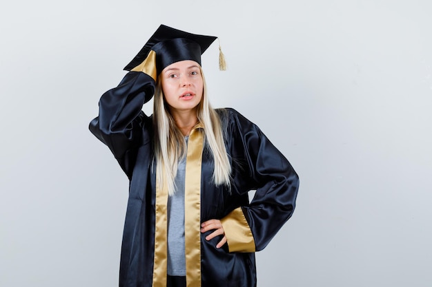 Blonde girl in graduation gown and cap holding hand on waist, putting hand on head and looking confident