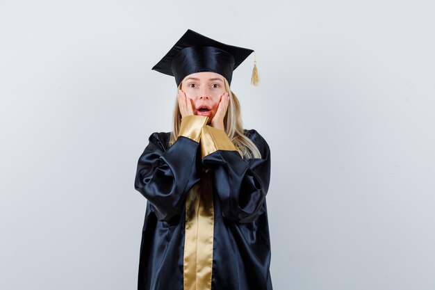 Blonde girl in graduation gown and cap holding hands on cheeks, opening mouth and looking surprised