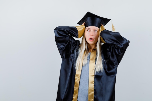 Blonde girl in graduation gown and cap holding hands on temples, looking at left side and looking surprised