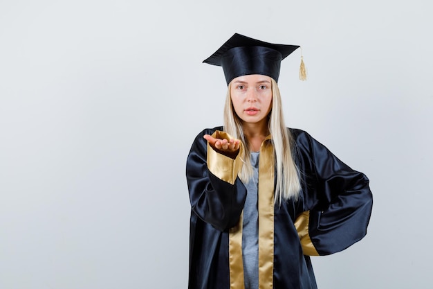 Blonde girl in graduation gown and cap holding one hand on waist, stretching another hand as holding something and looking charming