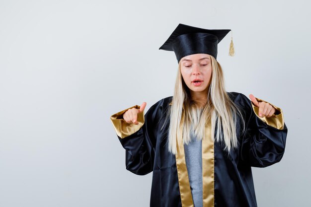 Blonde girl in graduation gown and cap pointing at camera with index fingers and looking serious