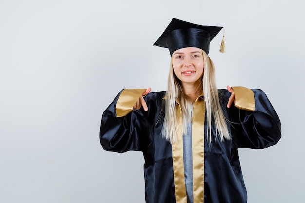 Blonde girl in graduation gown and cap pointing down with index fingers and looking cute