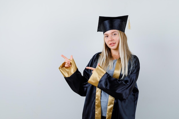 Free photo blonde girl in graduation gown and cap pointing left with index fingers and looking cute