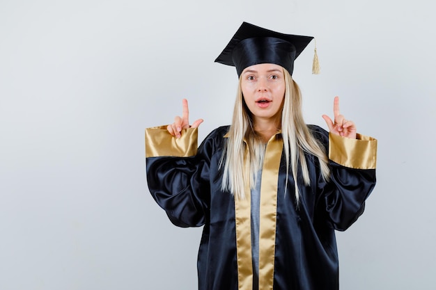 Blonde girl in graduation gown and cap pointing up with index fingers and looking cute