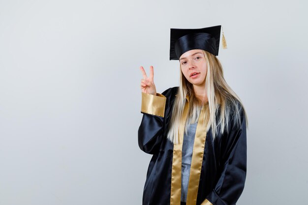 Blonde girl in graduation gown and cap showing peace gesture and looking cute