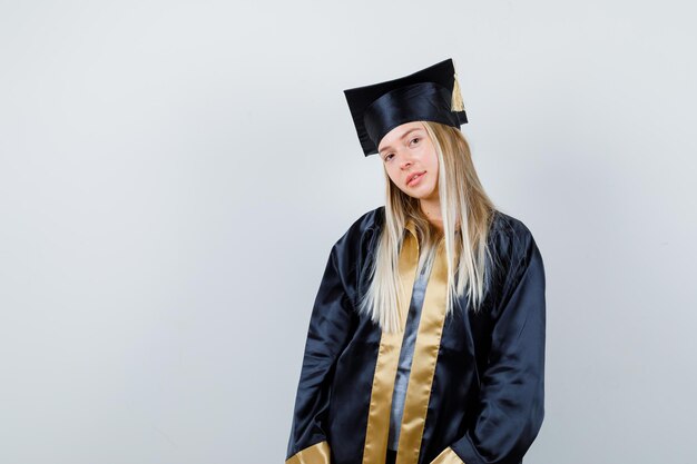 Blonde girl in graduation gown and cap standing straight and posing at camera and looking cute