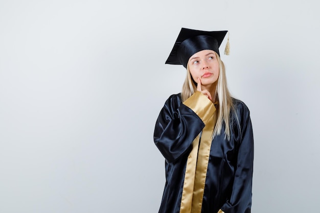 Blonde girl in graduation gown and cap standing in thinking pose and looking pensive