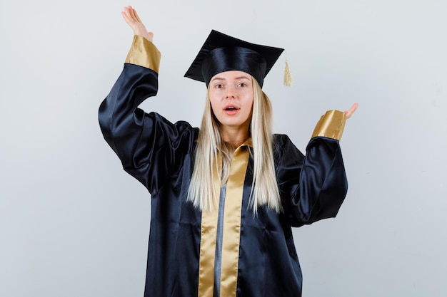 Blonde girl in graduation gown and cap stretching hands in questioning manner and looking happy